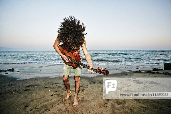 Gemischtrassiger Mann  der sich die Haare wirft und am Strand Gitarre spielt