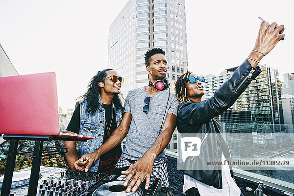 Men posing for cell phone selfie with DJ on urban rooftop