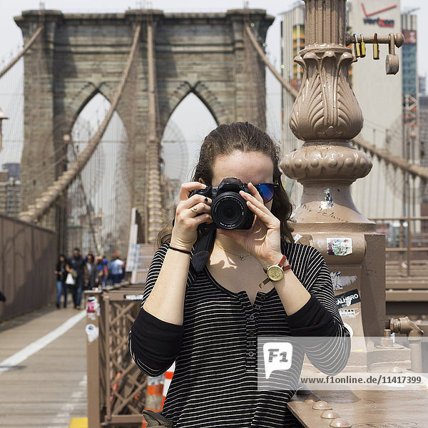 'A young woman holds a camera to take a photograph  pointing at the camera  on the Brooklyn Bridge; New York City  New York  United States of America'