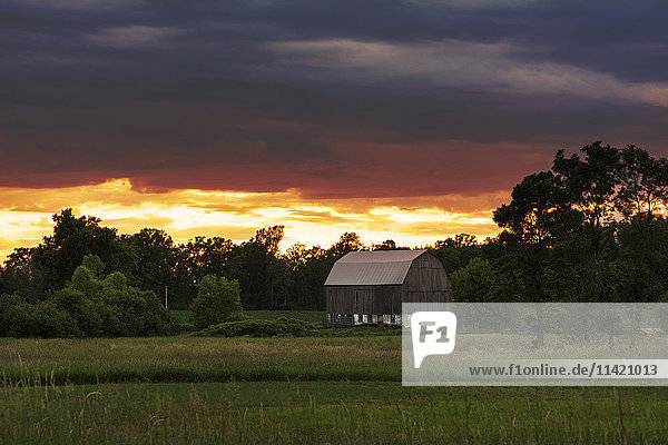 Ein roter Himmel bei Sonnenuntergang bildet einen farbenfrohen Hintergrund für ein graues Farmgebäude; Ontario  Kanada'.