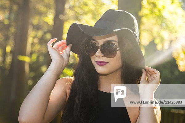 Ein fröhliches und modebewusstes Teenager-Mädchen posiert vor der Sonne mit einer Sonnenbrille und einem Designer-Hut  um ihre blassen Schultern zu schützen  während sie ihren Hut zum Schutz vor der Sonne hält; Vancouver  British Columbia  Kanada'.