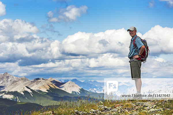 'Male hiker standing on top of rocky hill overlooking mountain range and valley with blue sky and clouds  West of Bragg Creek; Alberta  Canada '