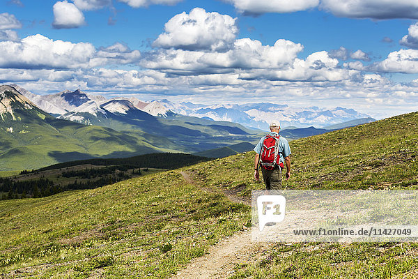 'Male hiker walking along a hillside trail overlooking mountain range and valley with blue sky and cloud  West of Bragg Creek; Alberta  Canada'