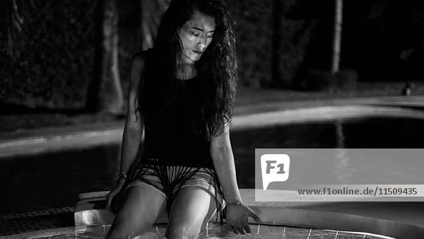 Junge erwachsene Frau sitzt am Rande eines Swimmingpools mit schimmerndem Wasser  das sich auf ihrem Körper spiegelt
