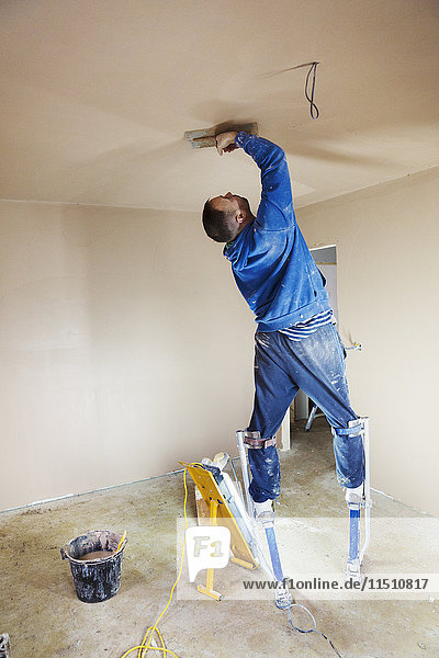 Ein Stuckateur mit Stelzen  der hoch oben an den Wänden eines im Bau befindlichen Hauses frischen Putz glättet.