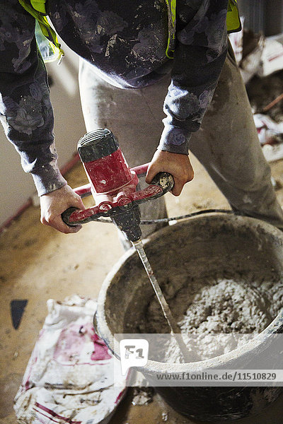 Ein Bauarbeiter  der auf einer Baustelle Putz mit einem elektrischen Rührwerk in einem Eimer mischt.