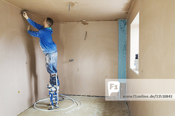 Ein Stuckateur mit Stelzen  der hoch oben an den Wänden eines im Bau befindlichen Hauses frischen Putz glättet.