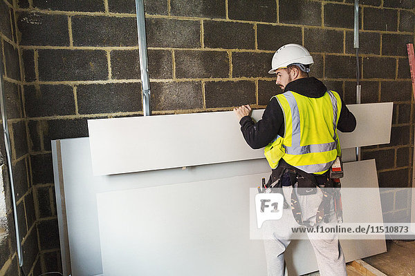 Ein Arbeiter mit Schutzhelm und High-Vis-Jacke  der auf einer Baustelle Holz gegen eine Wand stapelt.