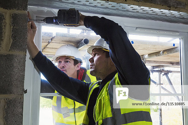 Zwei Arbeiter auf einer Baustelle  Bauarbeiter mit Schutzhelm  die eine elektrische Bohrmaschine an einem Fensterrahmen benutzen.