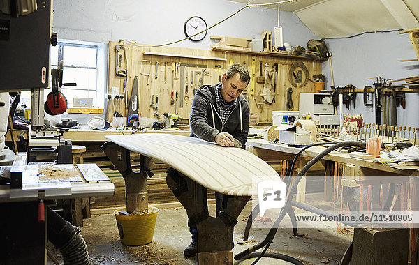 Mann steht in einer Werkstatt und schleift und formt ein hölzernes Surfbrett.