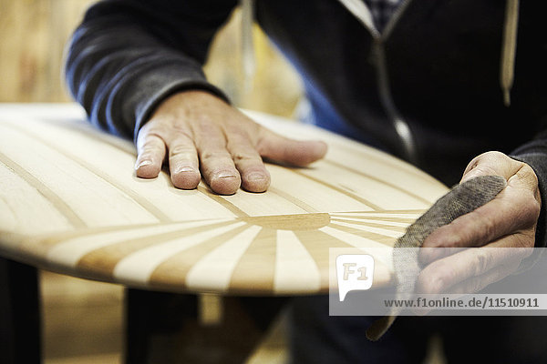 Mann in einer Werkstatt beim Schleifen der Kante eines hölzernen Surfbretts mit einem Oberflächenmuster aus eingelegtem Holz.
