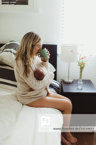 Mittlere erwachsene Frau sitzt im Bett und trinkt Kaffee  während sie ein neugeborenes Kind wiegt