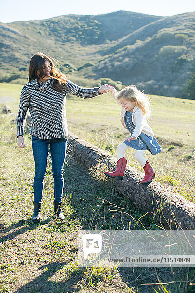Mittelgroße erwachsene Frau hält die Hand ihrer Tochter  während sie vom Baumstamm springt