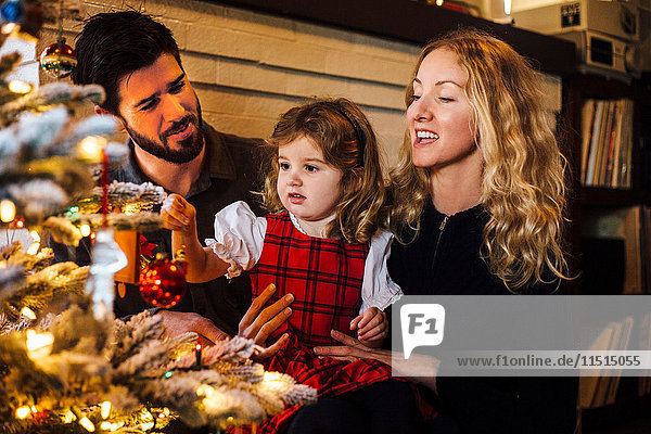 Weibliches Kleinkind mit Eltern  die eine Kugel auf den Weihnachtsbaum legen