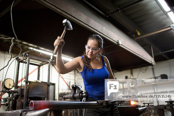 Female metalsmith hammering red hot metal rod on workshop anvil