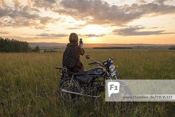 Kaukasische Frau mit Motorrad im Feld beim Fotografieren des Sonnenuntergangs