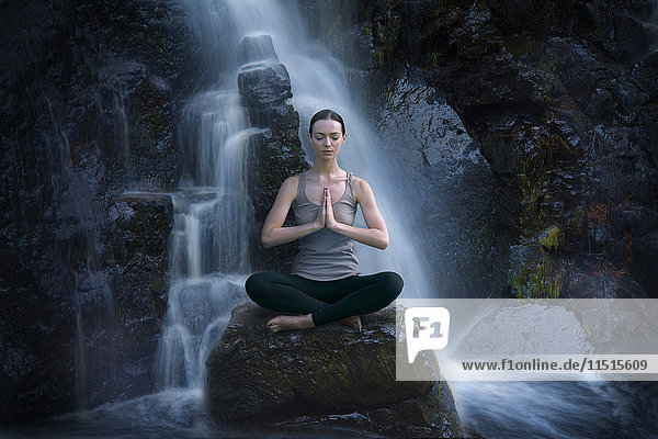 Kaukasische Frau meditiert auf einem Felsen in der Nähe eines Wasserfalls