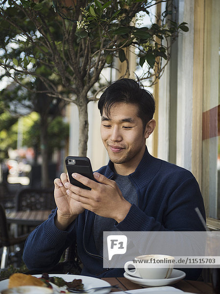 Lächelnder chinesischer Mann  der in einem Café im Freien eine SMS auf seinem Handy schreibt