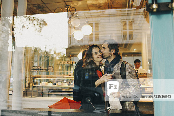 Caucasian man kissing woman on cheek behind bakery window