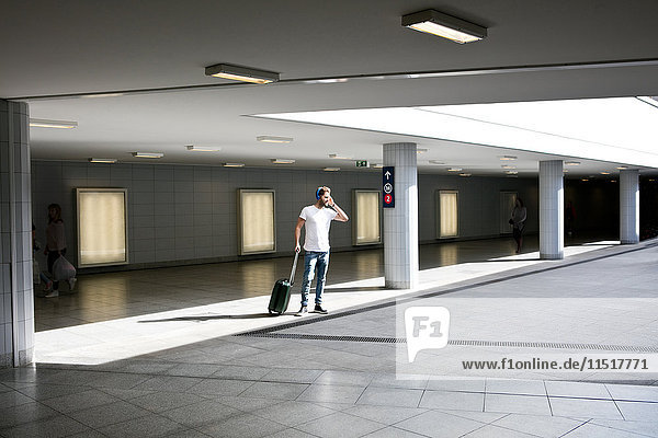 Young man in station  pulling wheeled suitcase  wearing headphones  using smartphone