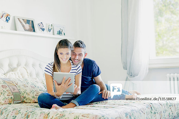 Ein schwangeres Paar sitzt auf dem Bett und benutzt ein digitales Tablet und lächelt