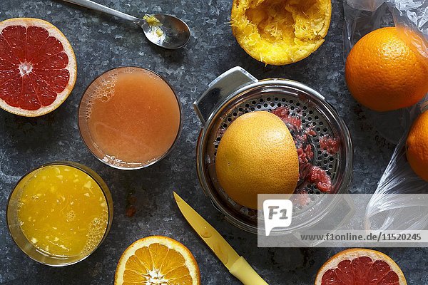 Manuelle Zitrusfruchtpresse aus Metall neben Gläsern mit Orangensaft und rotem Grapefruitsaft sowie ganzen und halbierten Orangen und roten Grapefruits