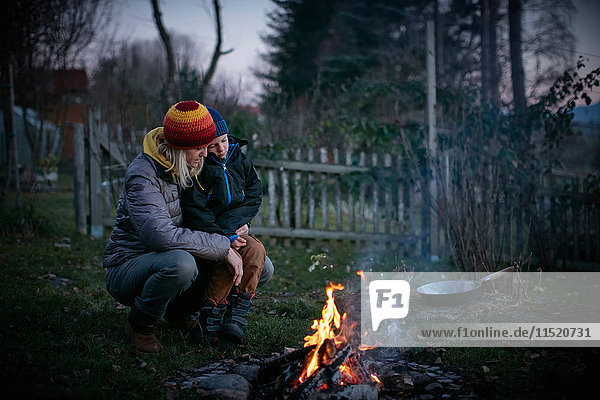 Mature woman and son crouching to watch garden campfire at dusk