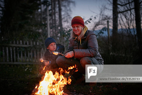 Reife Frau und Sohn rösten in der Abenddämmerung Marshmallows am Gartenlagerfeuer