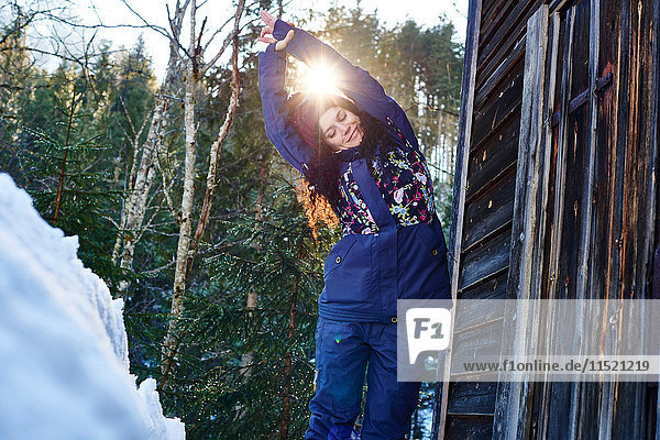 Frau in Winterkleidung übt Halbmond-Yoga-Pose im Schnee bei Blockhaus  Österreich