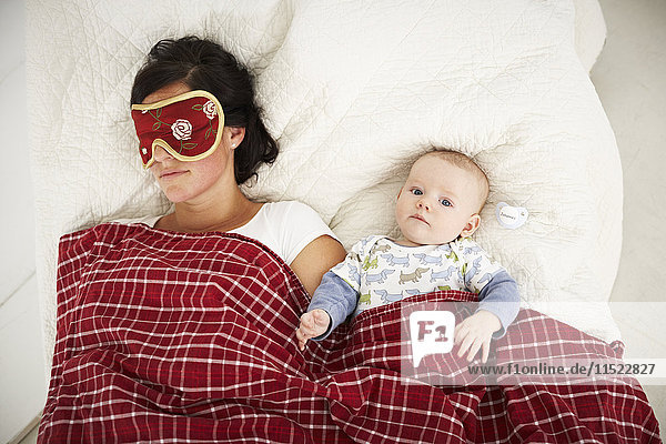 Mutter mit Schlafmaske und wachem Baby im Bett liegend
