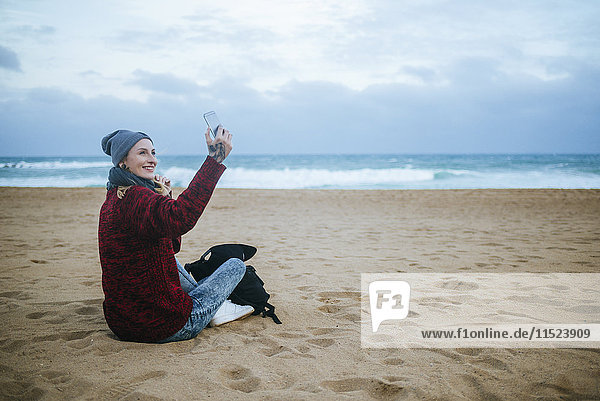 Lächelnde junge Frau  die im Winter am Strand sitzt und ihr Handy benutzt.