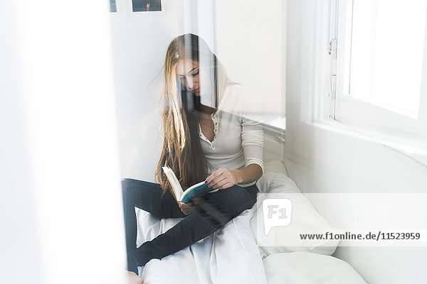 Young woman sitting on bed reading book