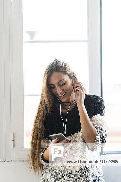 Lächelnde junge Frau mit Handy und Kopfhörer am Fenster