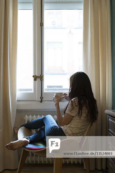 Woman with cup of coffee sitting on chair at home looking through window