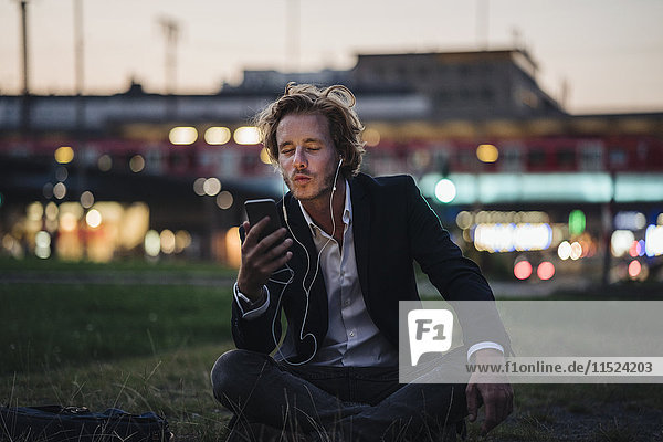 Businessman sitting on meadow at dusk with cell phone and earphones