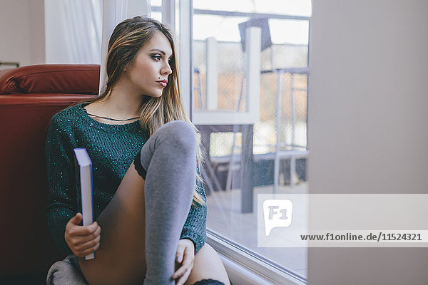 Junge Frau mit Buch sitzt auf dem Boden des Wohnzimmers und schaut durch die Balkontür.