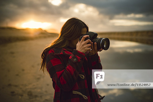 Junge Frau beim Fotografieren am Strand mit Kamera bei Sonnenuntergang