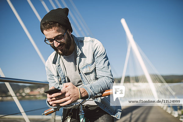 Smiling young man with fixie bike using a smartphone on a bridge