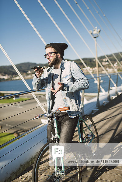 Junger Mann mit Fixie Bike auf einer Brücke mit Smartphone