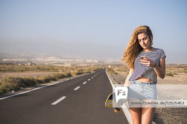 Spanien  Teneriffa  junge Frau mit Skateboard auf leerer Landstraße mit Blick auf Smartphone