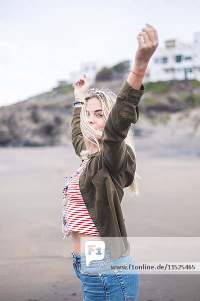 Porträt einer blonden jungen Frau am Strand mit erhobenen Armen