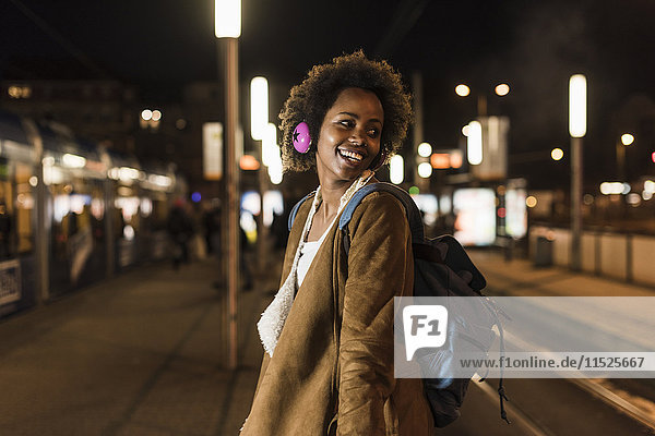 Smiling young woman with headphones and backpack waiting at the tram stop