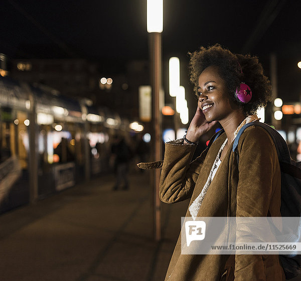 Smiling young woman listening music with headphones while waiting at the tram stop
