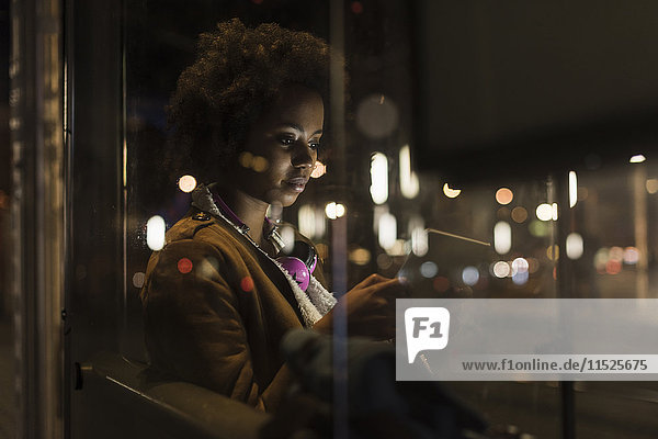 Junge Frau mit Tablette wartet an der Straßenbahnhaltestelle bei Nacht
