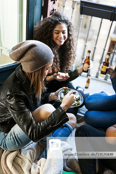 Gruppe von Freunden sitzt auf dem Boden und isst Pizza und Salat und trinkt Bier zu Hause.