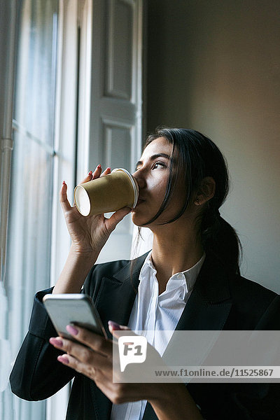 Junge Geschäftsfrau am Fenster  die Kaffee trinkt und Handy hält