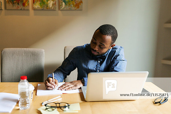 Junger Mann am Tisch mit Laptop-Schriftzug im Notizbuch