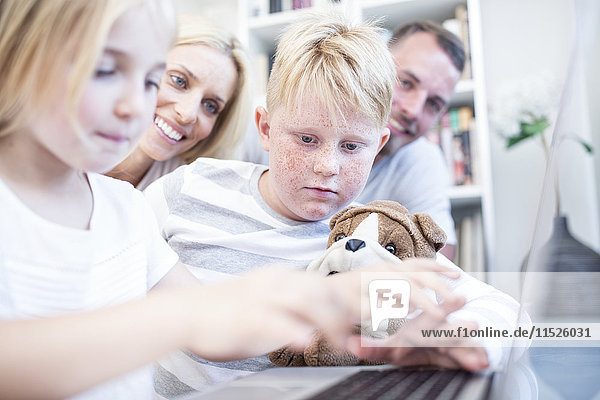 Brother and sister with cuddly toy using laptop together watched by parents