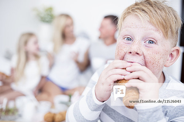 Porträt eines Jungen  der in ein Croissant beißt  mit Familie im Hintergrund