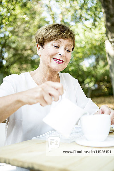 Lächelnde Seniorin gießt Milch in die Tasse Kaffee im Freien